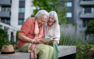 Elderly mother sitting on bench with adult daughter, reading together.